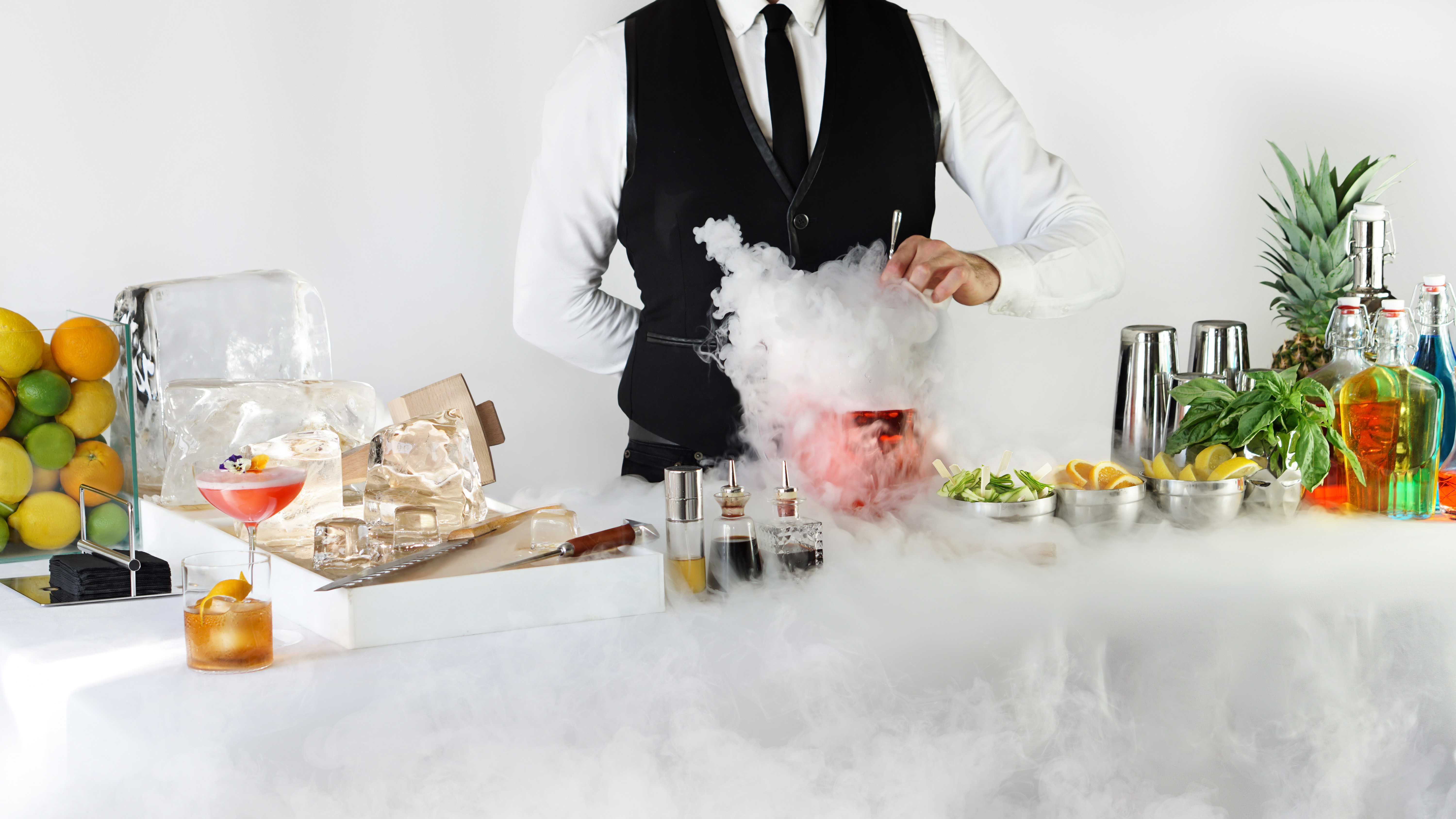 White bar with ice and fruits on display and a bartender making red cocktail in a pitcher with dry ice