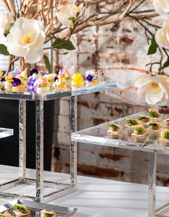 Event food station with colorful hors d'oeuvres on acrylic platters on stainless steel elevations and magnolia flowers in background
